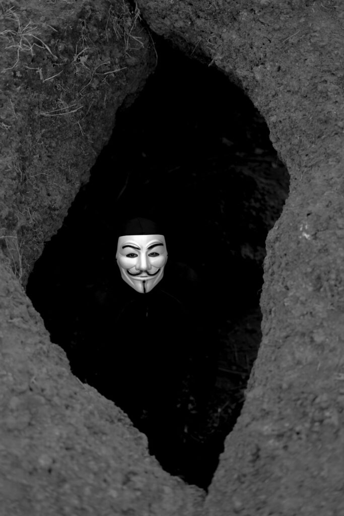 A person wears an anonymous mask, peeking out from a dark, eerie tunnel crevasse in grayscale.