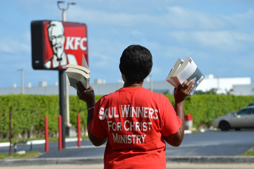 A street preacher in a red t-shirt holds books outside a KFC, advancing their message.
